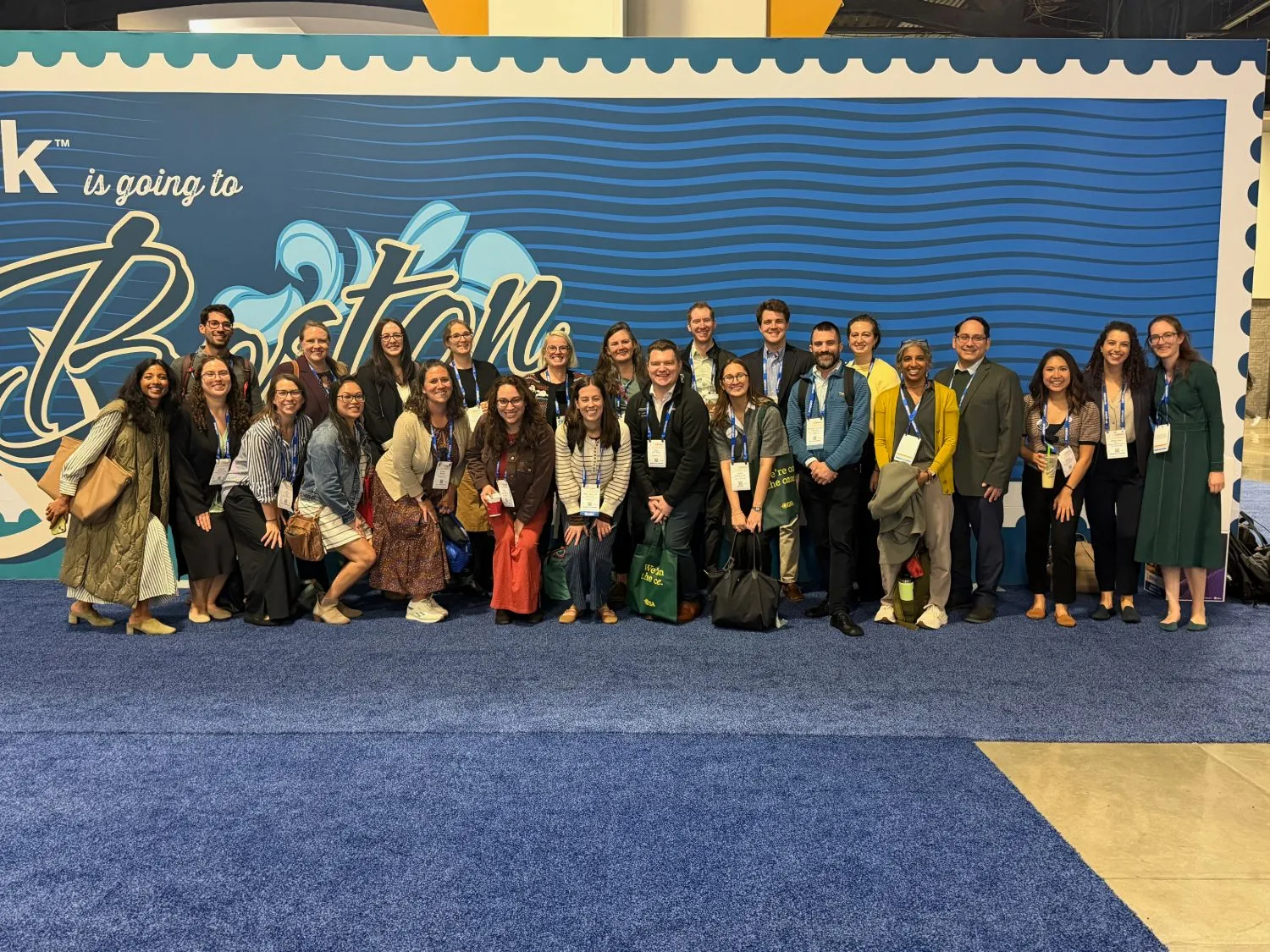 About two dozen people wearing IDWeek lanyards pose together in front of a large blue banner reading IDWeek is going to Boston.